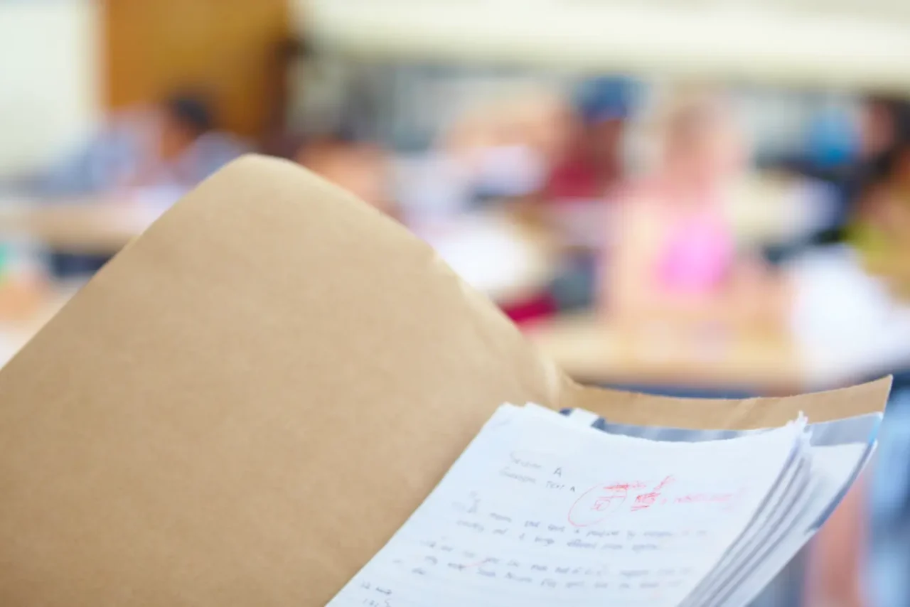 Close-up of lecture notes inside a brown folder, showing handwritten answers and teacher's red-ink feedback, with students in a blurred classroom background