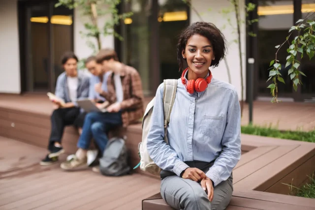 Smiling woman in a shirt sitting on a bench with red headphones and a backpack in the university courtyard.